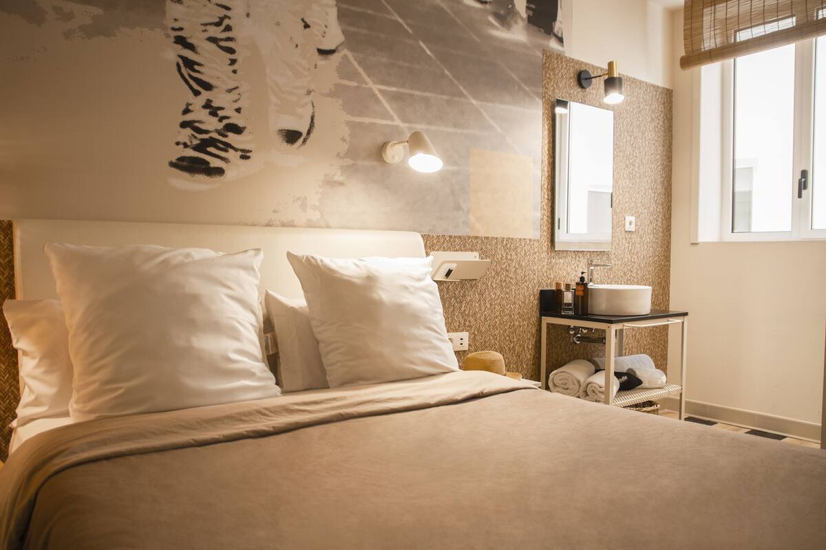 A bedroom with a bed, pillows, a bedside table with a lamp, a mirror, and a window.