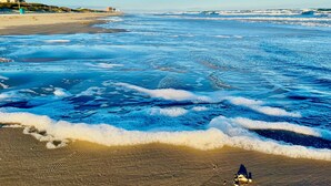 Ein Strand mit brechenden Wellen und feinem Sand, sowie ein Gebäude im Hintergrund.