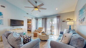 Ein modernes Wohnzimmer mit grauen Sofas, einem Couchtisch und einem Fernseher. Ein Deckenventilator, eine Tür zu einem Balkon und Kunstwerke an den Wänden sind ebenfalls vorhanden.