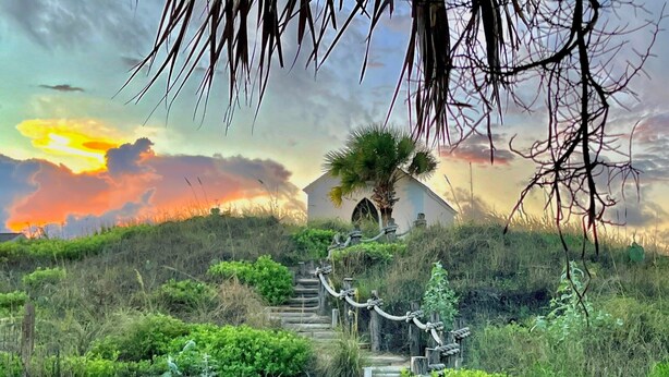 Ein Weg führt zu einem weißen Gebäude mit einer Palme, umgeben von üppiger Vegetation und einem Sonnenuntergang im Hintergrund.
