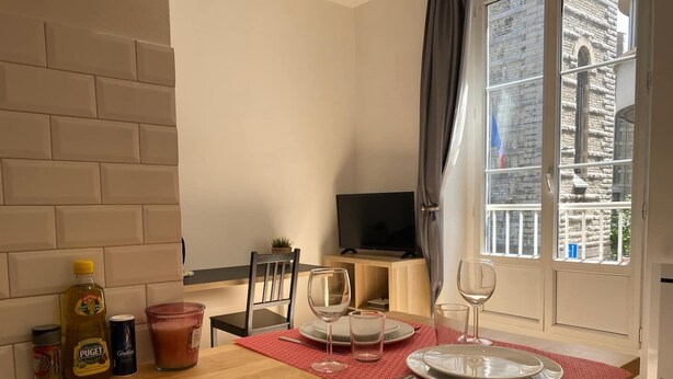 A dining area with a wooden table set for two, a TV, and a view of a stone building through a window. - Le Cocon / Hyper-Centre / Air-conditioning / Trams (Grenoble)