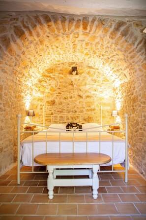 A bedroom with a stone wall, a bed with white bedding, two bedside lamps, and a wooden bench in front of the bed.