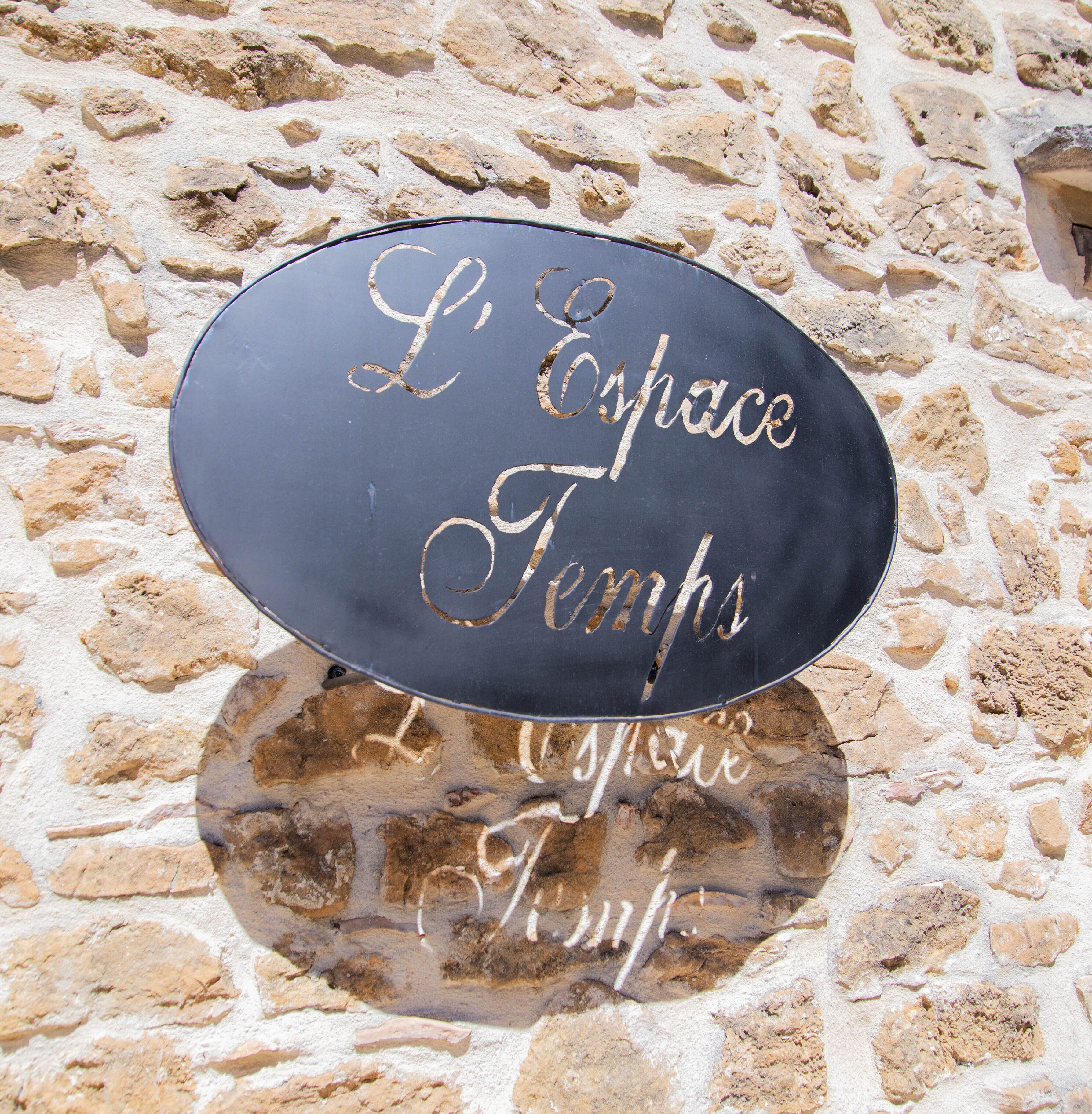 Un mur de pierre sur lequel est fixée une plaque noire réfléchissante gravée des mots « L’Espace Temps ».