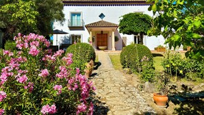 Un edificio blanco con techo de tejas, un camino de piedra que lleva a la entrada, y plantas con flores rosadas en primer plano.