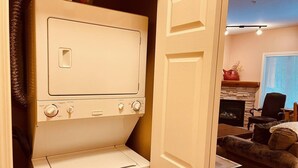 Un lavadero pequeño con lavadora y secadora, una puerta de madera, y una sala de estar con chimenea y asientos.