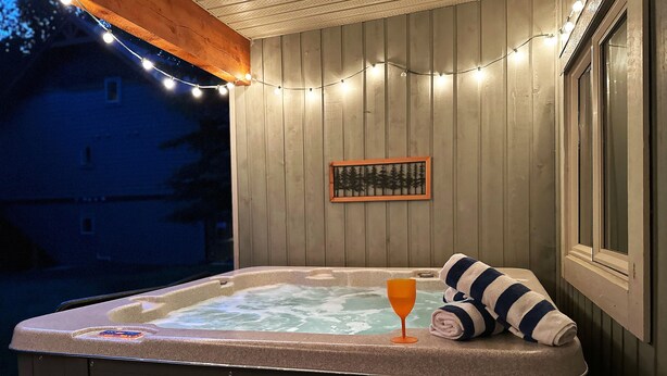 A hot tub with a towel and glass, surrounded by string lights, wooden walls, and a framed picture. - Willow Vistas 6 - Golf - Ski - Hike - Chill - Private Hot Tub - Sleeps 6 (Kimberley)