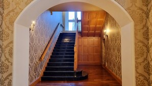 Un grand escalier en bois, doté d’une rampe, mène à une pièce dont les murs sont recouverts d’un papier peint à motifs et qui possède une porte en bois.