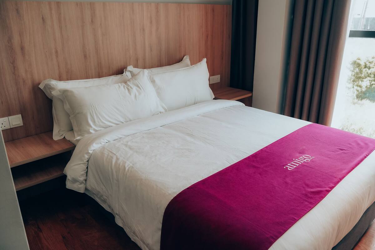 Un lit bien fait, avec des draps blancs et une couverture magenta, une tête de lit en bois et une fenêtre avec des rideaux.