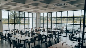 Un restaurante amplio con grandes ventanales que dan a un aparcamiento y coches. Las mesas están ordenadas con sillas, y el interior presenta un diseño moderno con suelo de baldosas.