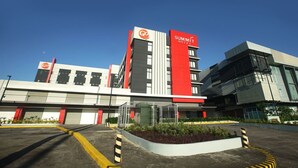 A modern hotel with a prominent red and white facade, multiple floors, and a parking area with yellow lines and bollards. - Go Hotels Plus Naga (Naga)