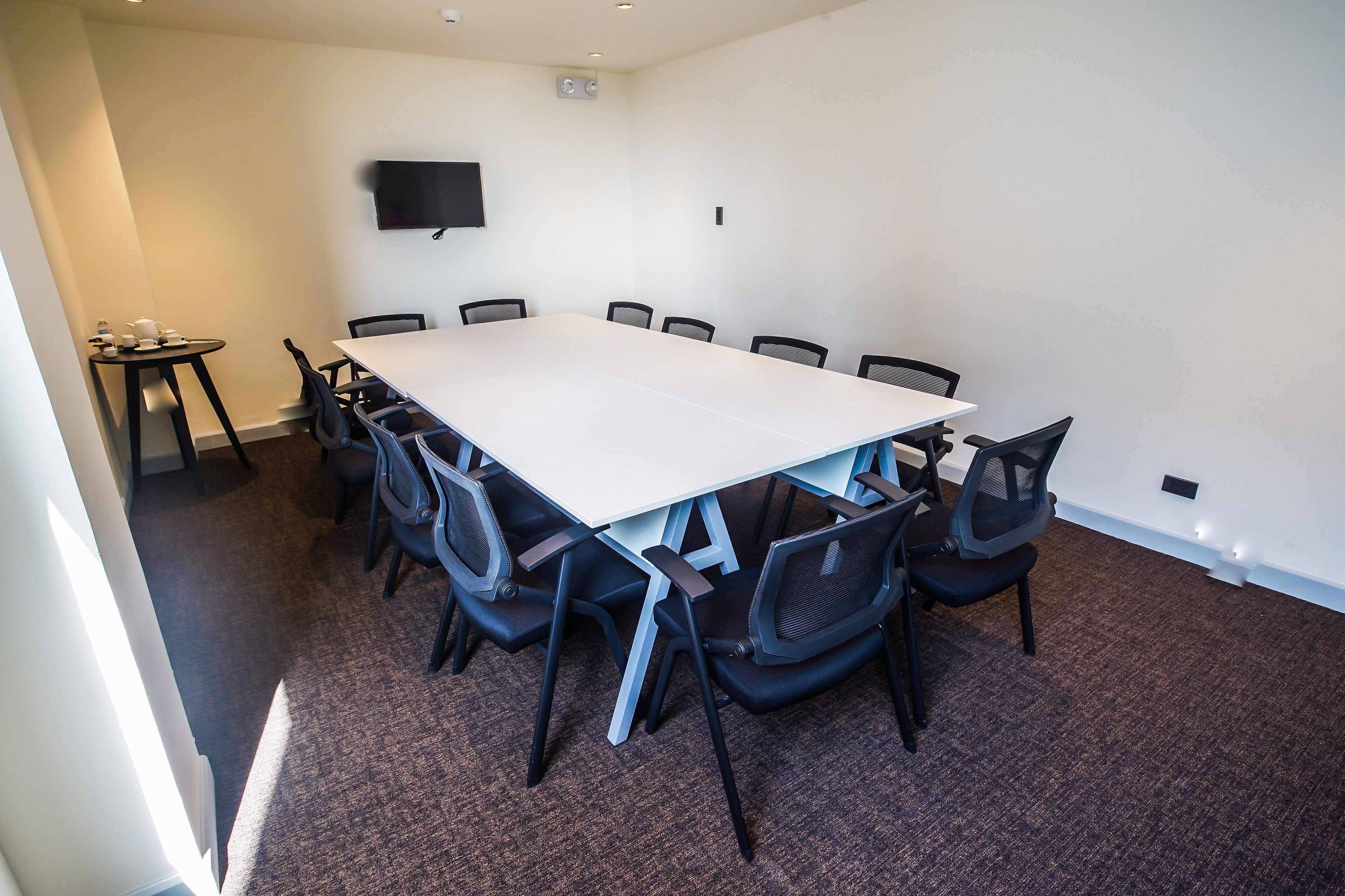 A conference room with a large rectangular table, black chairs, a small table with cups and a bottle, and a flat-screen TV mounted on the wall.