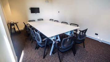 Uma sala de conferências com uma mesa retangular grande, cadeiras pretas, uma mesa pequena com copos e uma garrafa, e uma televisão de tela plana na parede.