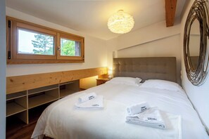 A bedroom with a large bed, two windows, a wooden bench, a bedside table with a lamp, and a decorative mirror.
