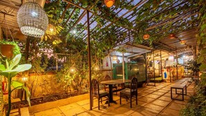 Un patio al aire libre con una mesa y sillas, rodeado de plantas en macetas e iluminación colgante.