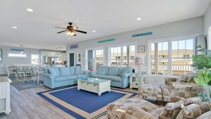 A spacious living room with a large window, a ceiling fan, and a view of the beach. - Rushin to the Beach: Spacious 8-bedroom oceanview home in Oak Island, perfect for families. (Oak Island)