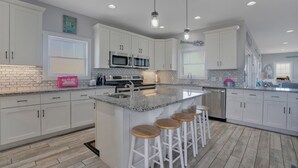 A modern kitchen with white cabinets, granite countertops, stainless steel appliances, and wooden stools. - Rushin to the Beach: Spacious 8-bedroom oceanview home in Oak Island, perfect for families. (Oak Island)