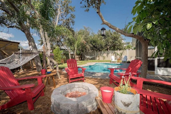 Un patio trasero con piscina, sillas Adirondack rojas, una hamaca y un pozo de fuego.