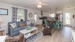 Een ruime woonkamer met een grijze bank, een salontafel en rieten stoelen. In de achtergrond is een keuken met een centraal eiland en barstoelen zichtbaar.