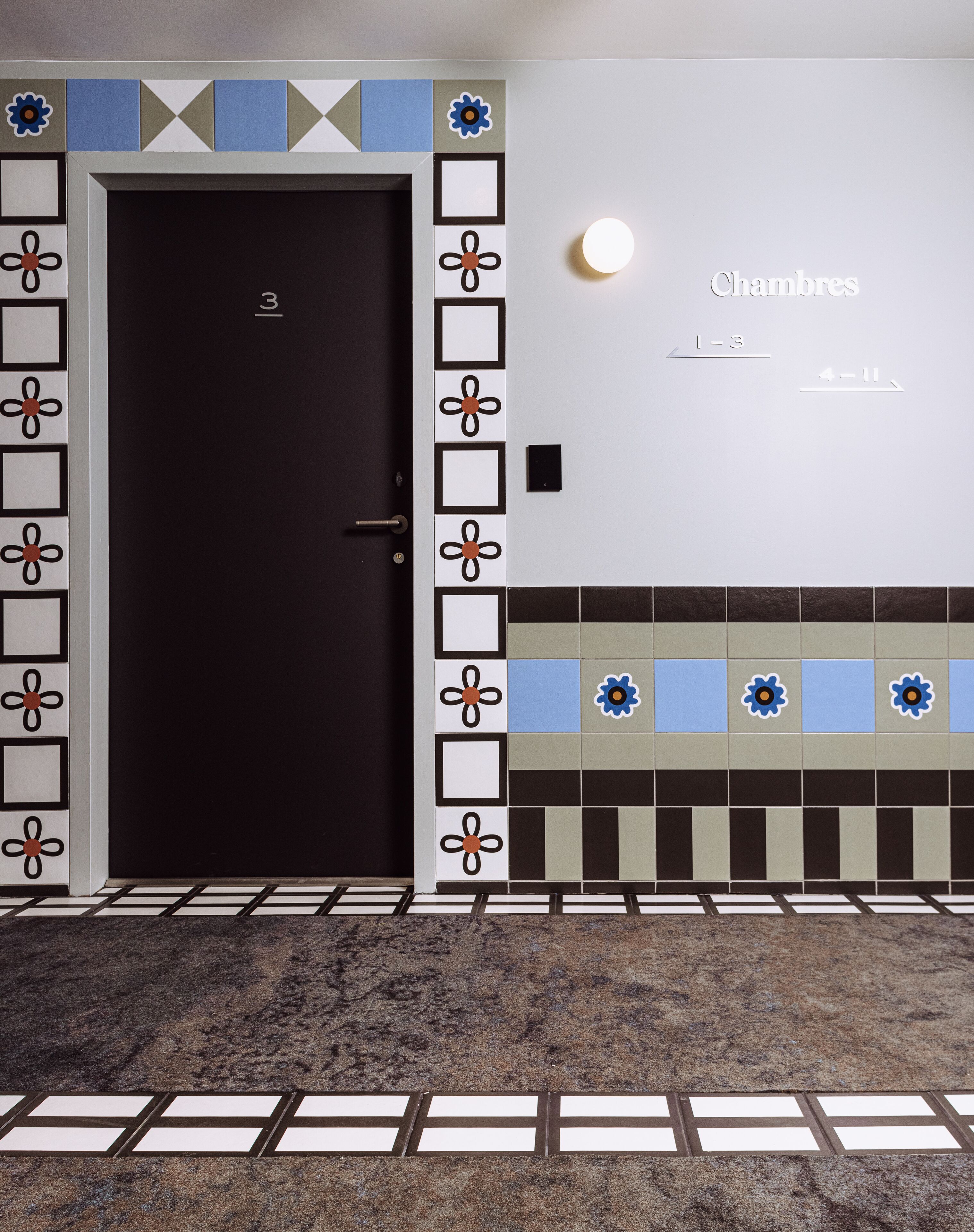Une chambre d’hôtel avec une porte sombre, une horloge murale et un mur recouvert de carreaux formant un motif géométrique.
