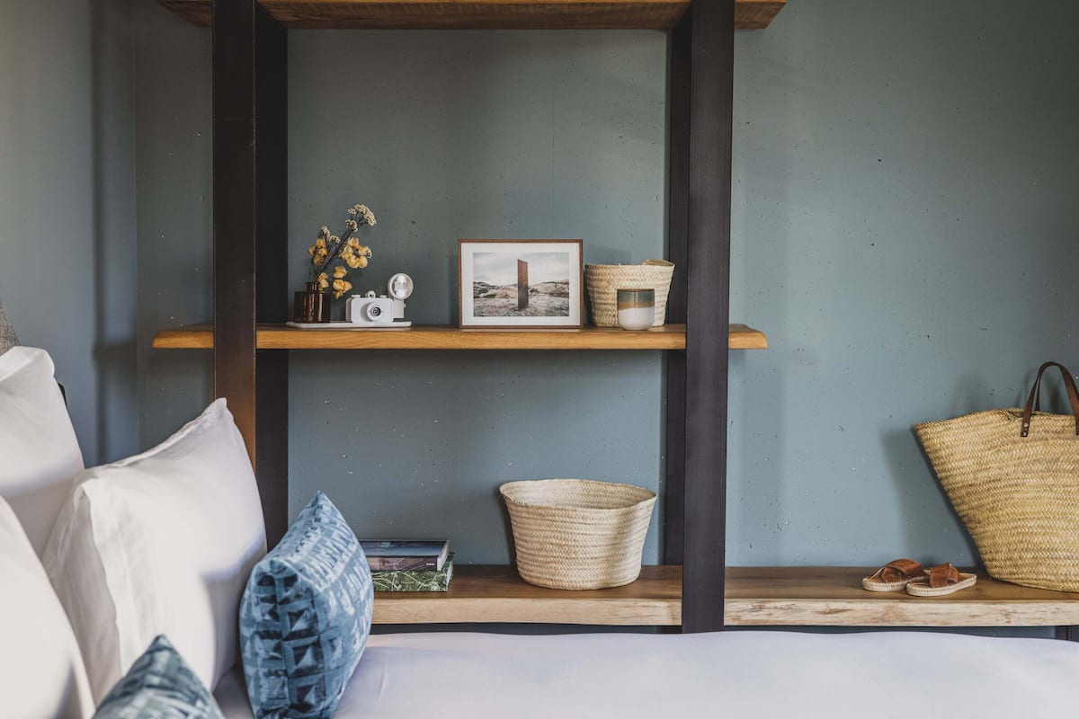 A bedroom with a wooden bed frame, a shelf with decorative items, and a large woven bag.