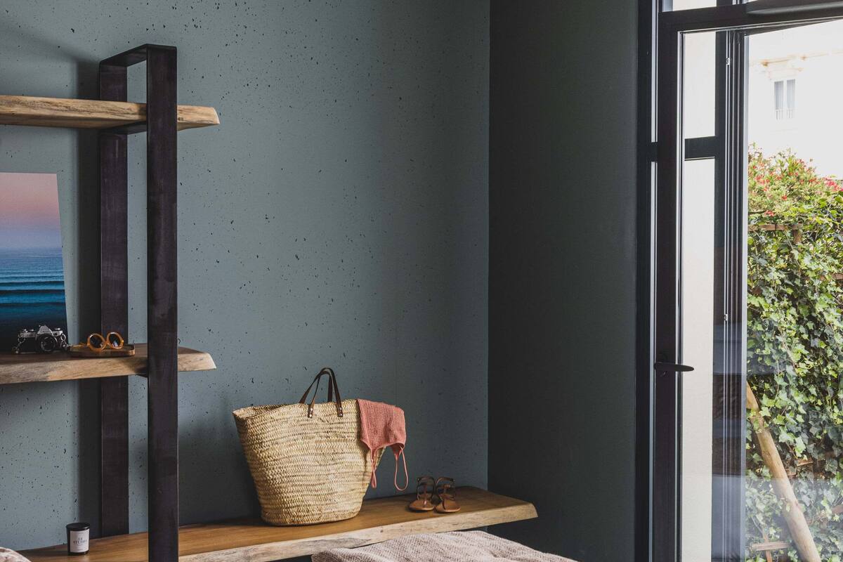 A bedroom with a bed, a wooden shelf with a basket, a painting, and a view of greenery through a window.