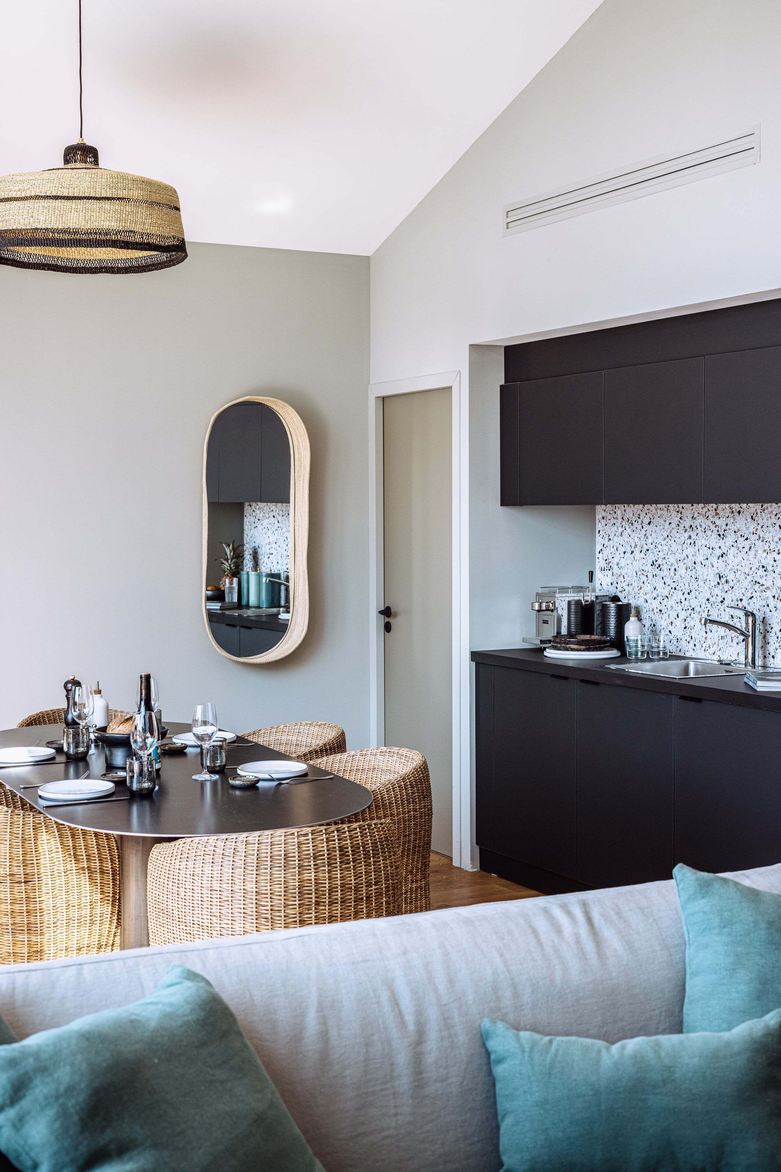 A modern kitchen with dark cabinets and a white backsplash, a dining area with wicker furniture and a round table, and a large mirror on the wall.
