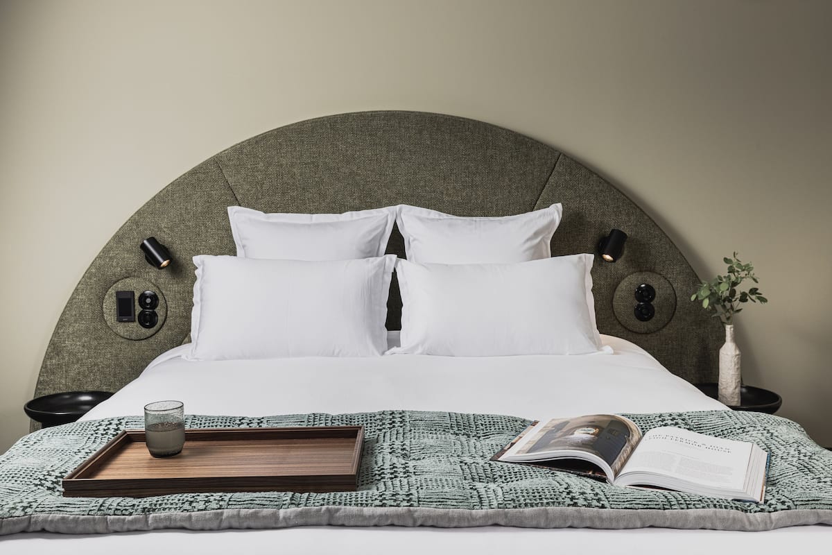 A neatly made bed with white linens, a tray with a glass and a book, and a bedside table with a vase and a lamp.