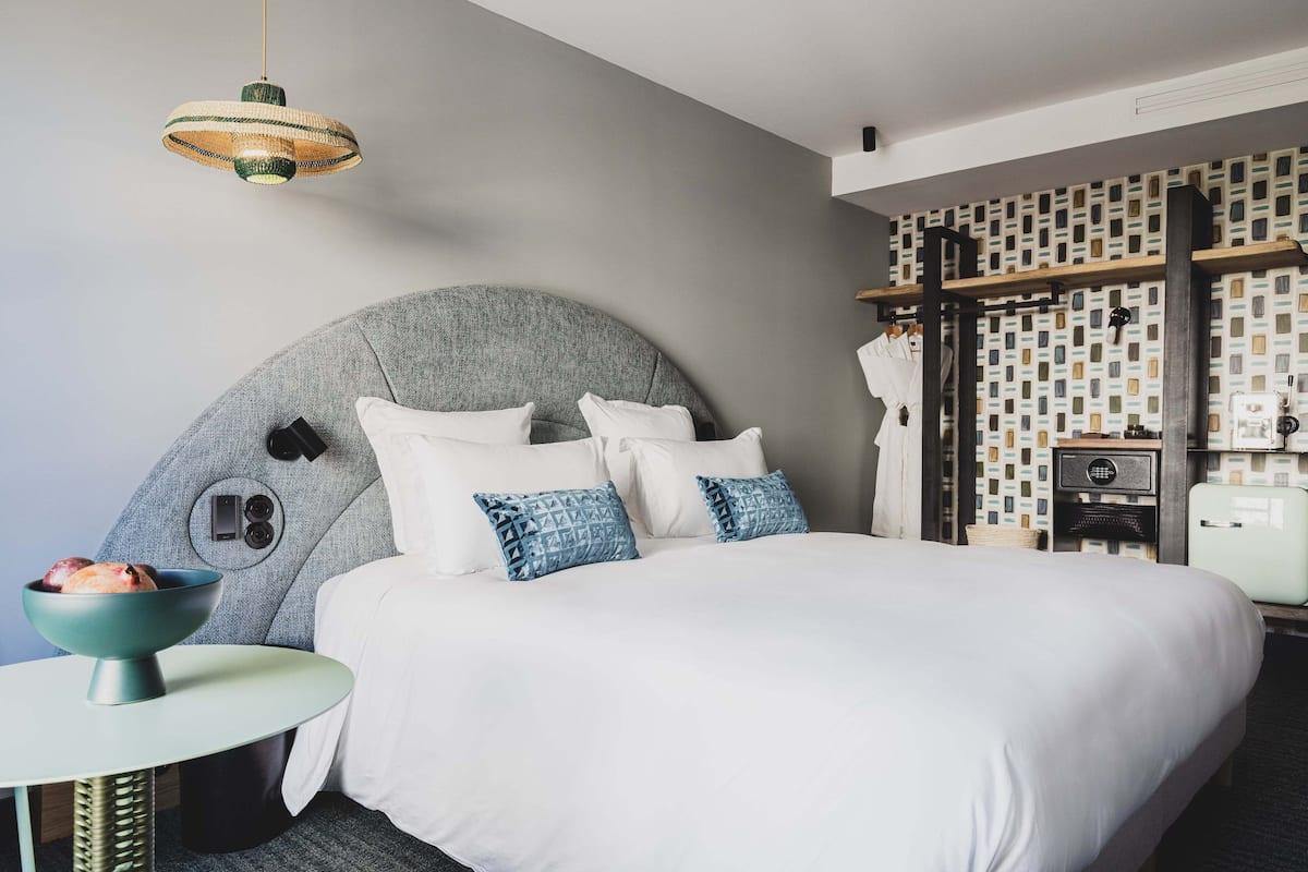 A modern hotel room with a large bed, a round table with a bowl of fruit, a unique wall design, and a hanging light fixture.