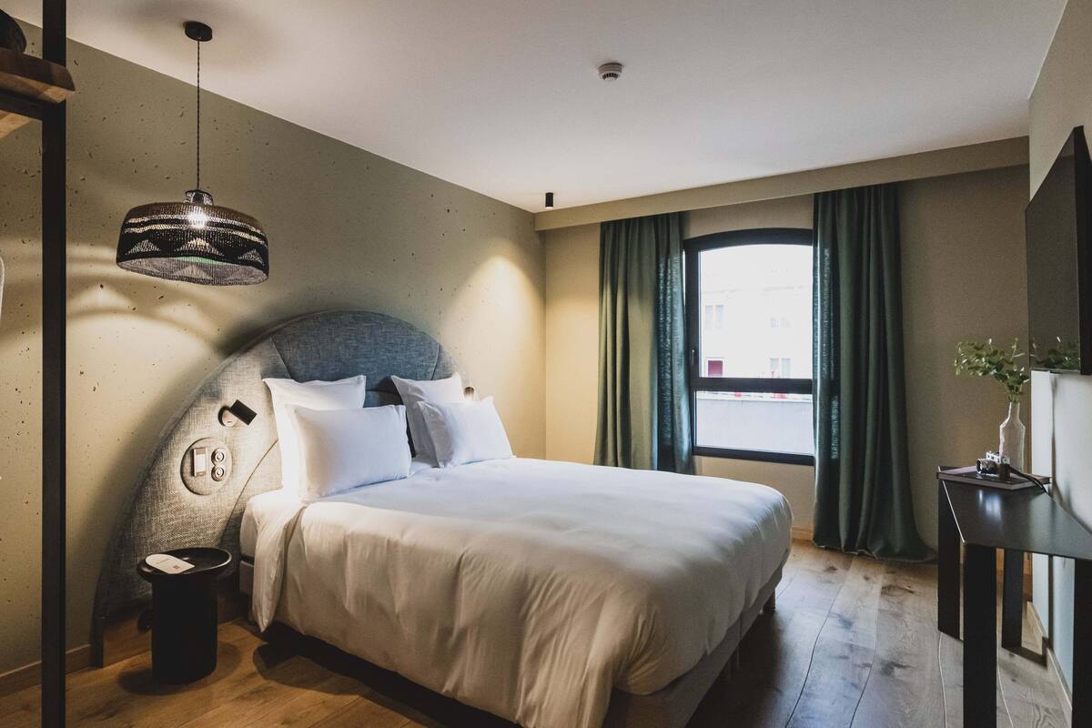 A bedroom with a bed, a hanging light fixture, a robe, a trash bin, and a window with curtains.