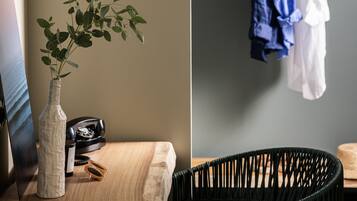 A room with a wooden table, a green woven lamp, a white vase with a plant, a black wicker chair, and a clothing rack with hanging clothes.