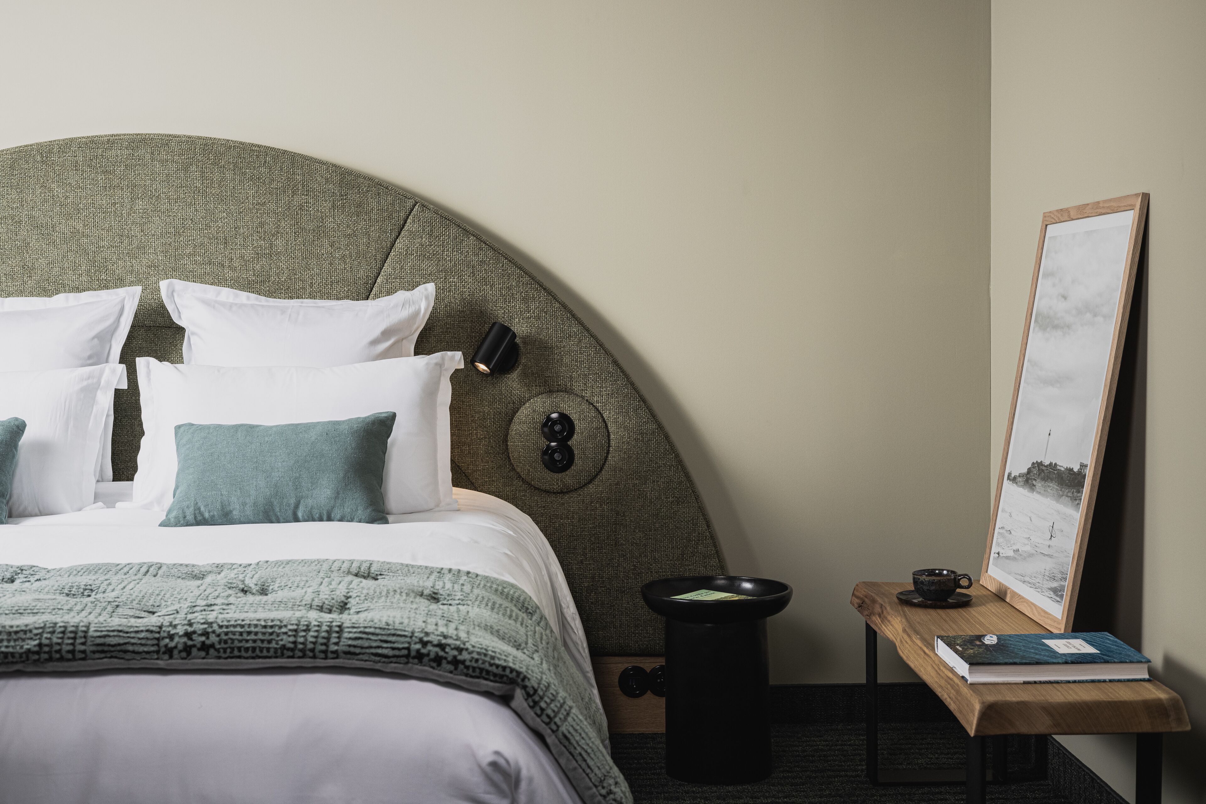 A neatly made bed with white and green bedding, a round headboard, a bedside table with a book and a cup, and a framed picture on the wall.