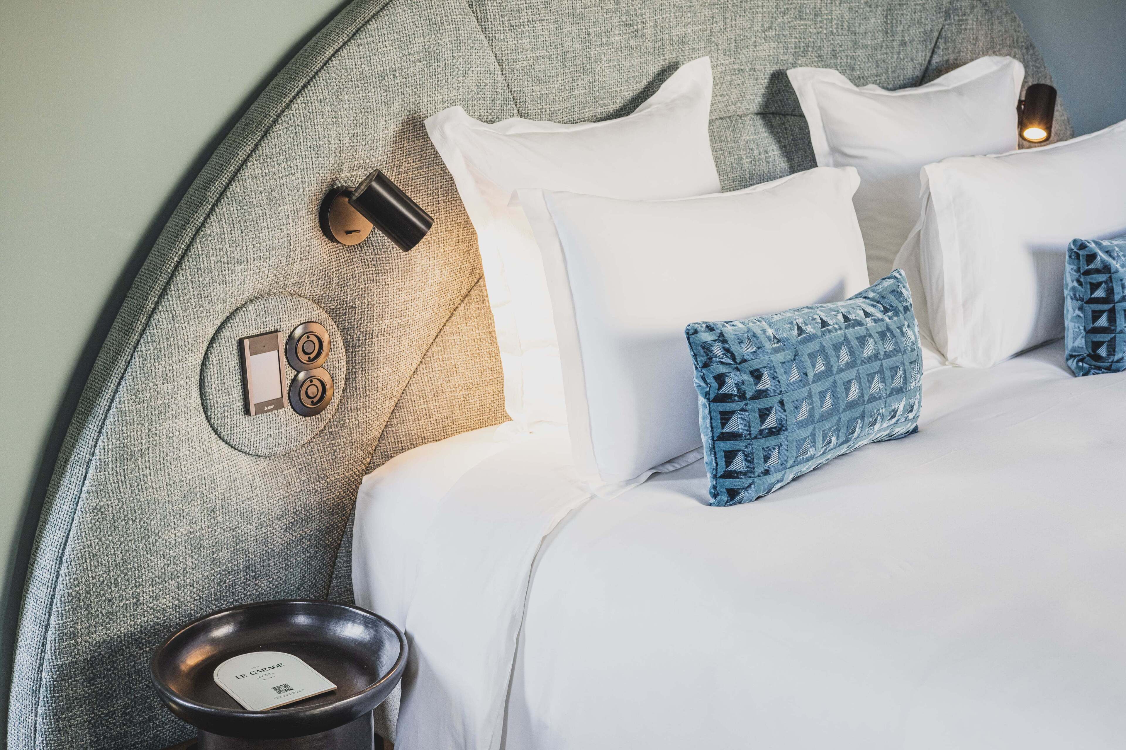 A neatly made bed with white linens and blue patterned pillows, a round bedside table with a book and a small lamp, and a textured headboard.