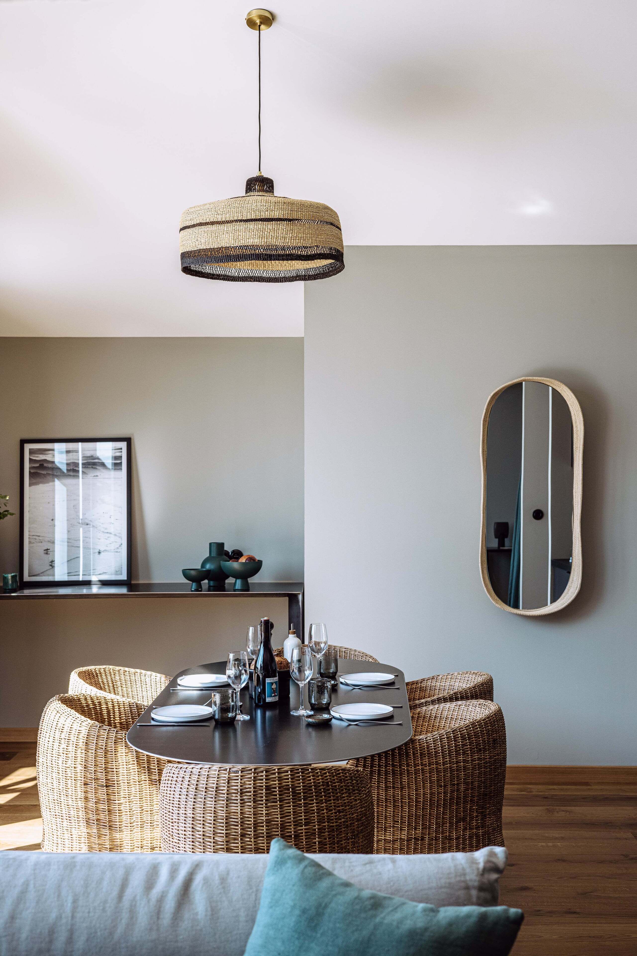 A dining area with a round table set for four, surrounded by wicker chairs, a hanging light fixture, and a wall-mounted mirror.