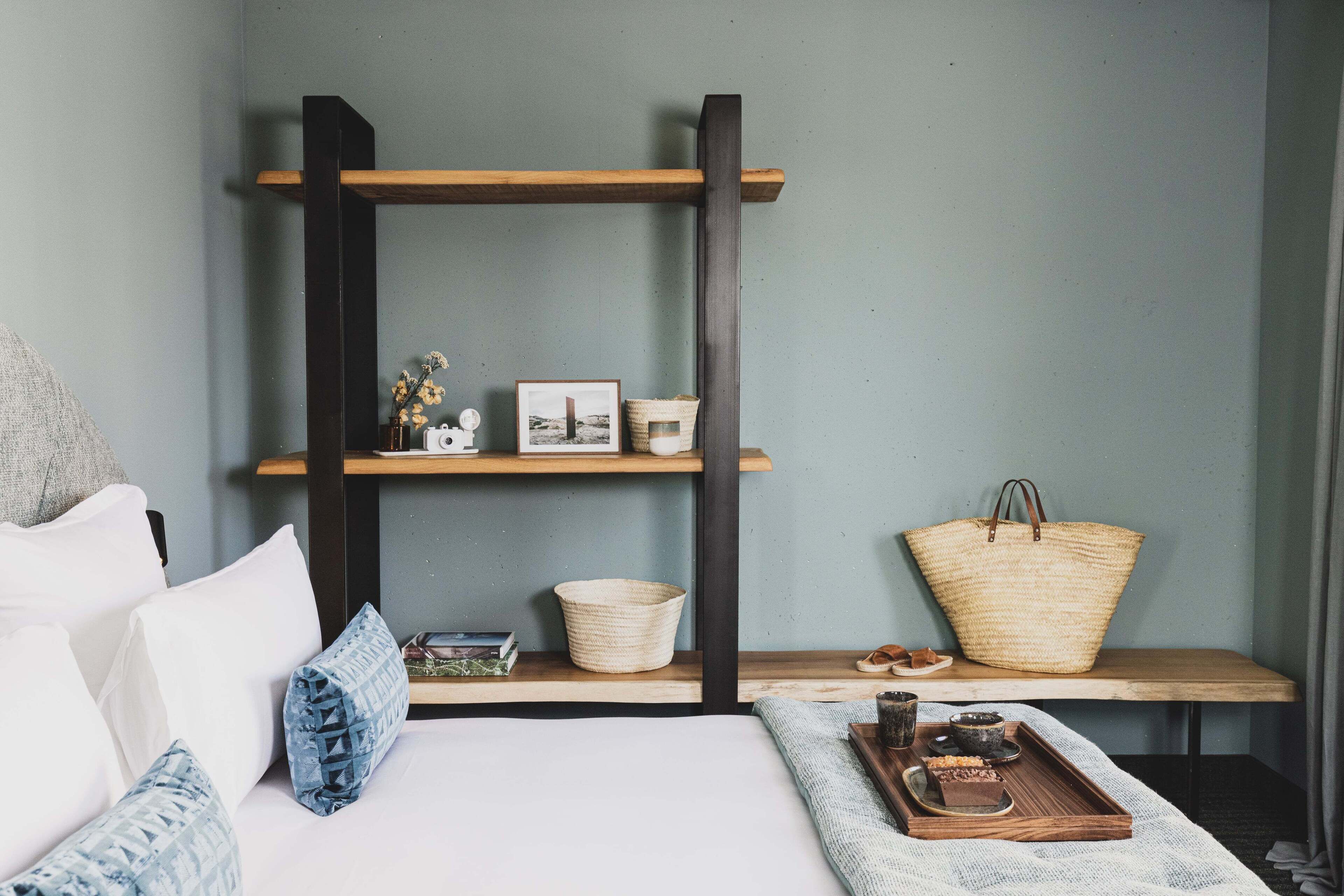 Une chambre à coucher bien rangée, avec un lit, des oreillers, une table de chevet sur laquelle se trouve un plateau, une étagère avec des objets décoratifs et un grand panier en osier.
