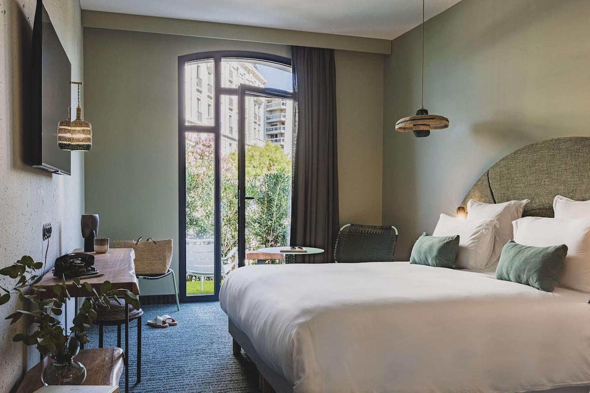 A hotel room with a large bed, a desk with a telephone, a chair, a small table, and a view of a building and greenery outside.