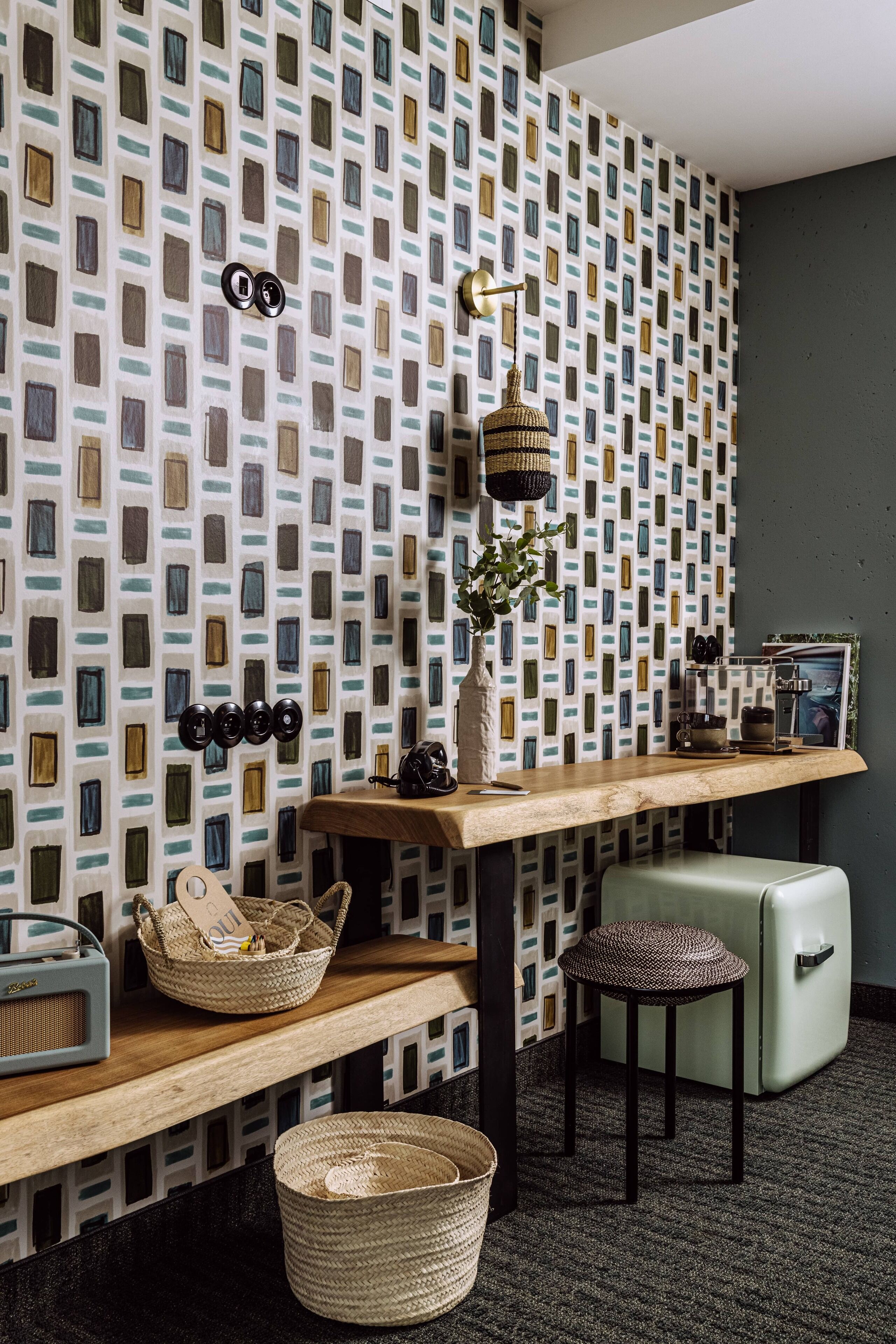 Ein Zimmer mit gemustertem Wandpapier, einem Holztisch, einem alten Radio, einem Korbwäscheskorb, einem Hocker und einem Vintage-Kühlschrank.