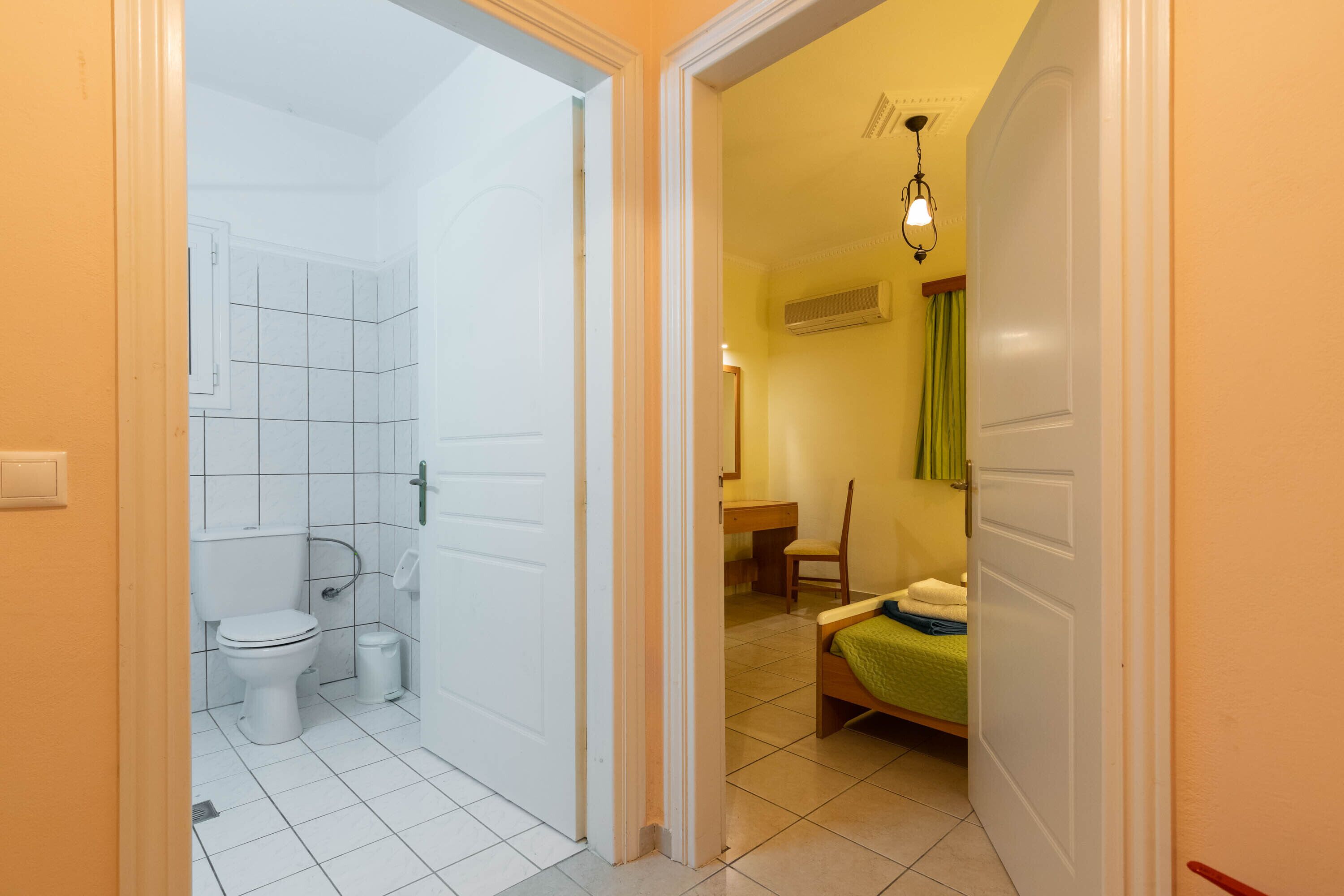 Un baño con inodoro, lavamanos y ducha. En la habitación contigua, hay una cama con una manta verde y una silla.