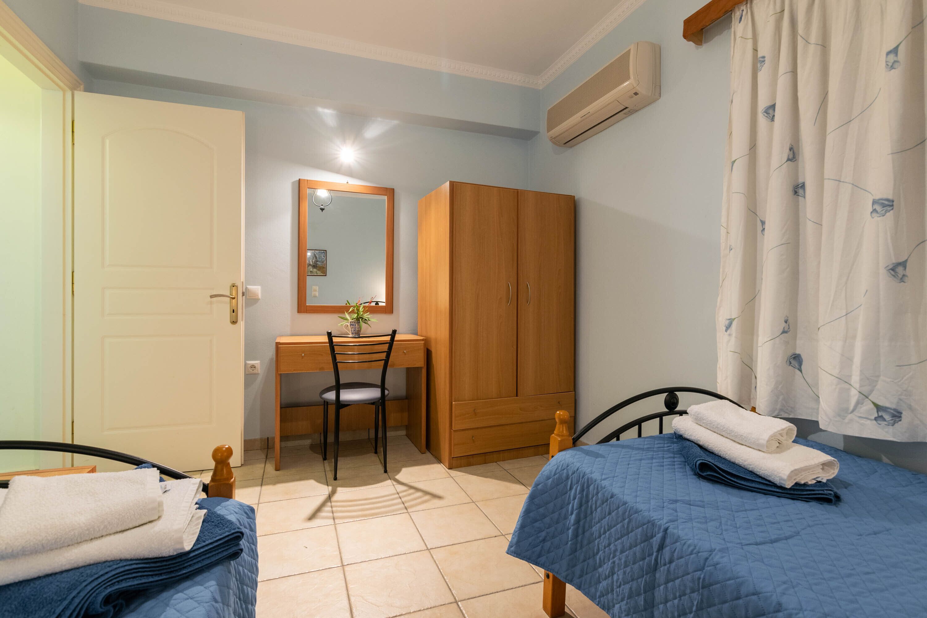 A bedroom with two beds, a desk, a chair, a wardrobe, and a bathroom with a shower curtain.