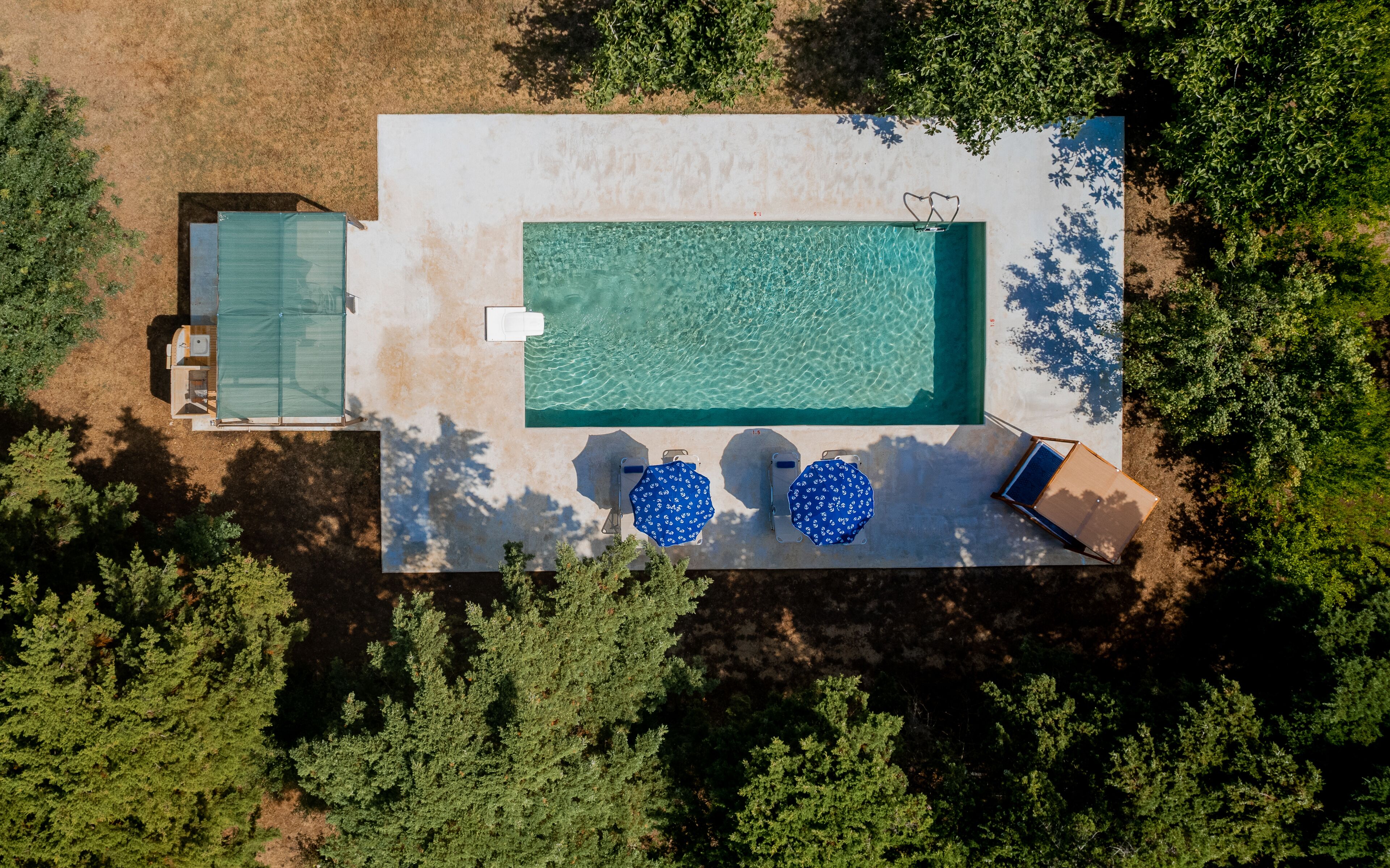 Vista aérea de una piscina rectangular con agua cristalina, rodeada por una zona de terraza con dos sombrillas azules y una tumbona.