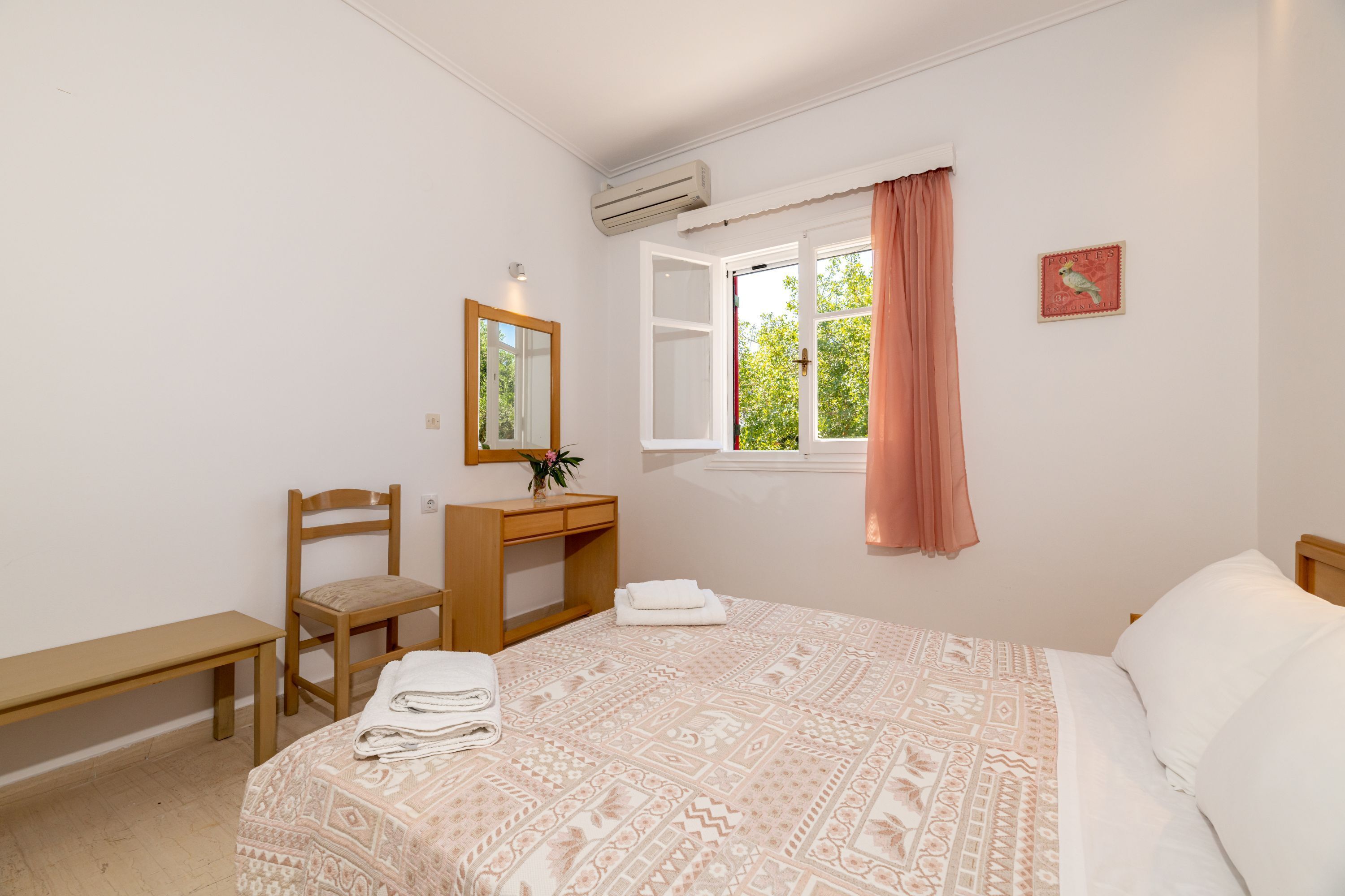 A bedroom with a bed, a desk, a chair, a bench, a window with curtains, and a wall-mounted air conditioner.