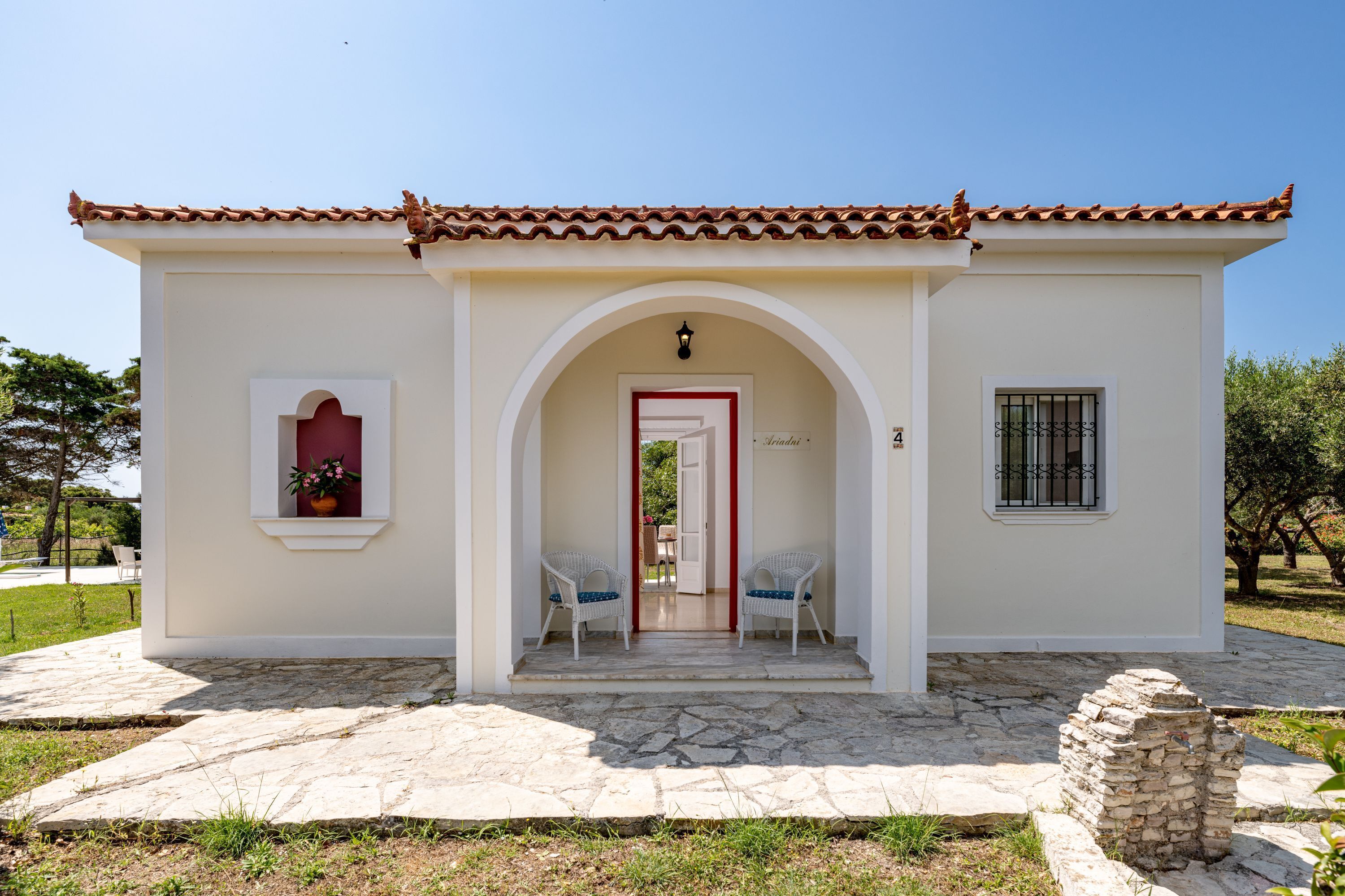 A small building with a tiled roof, a stone-paved entrance, and two white chairs outside.