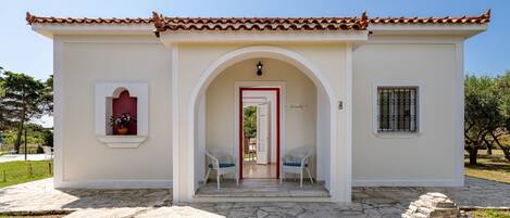 A small building with a tiled roof, a stone-paved entrance, and two white chairs outside.