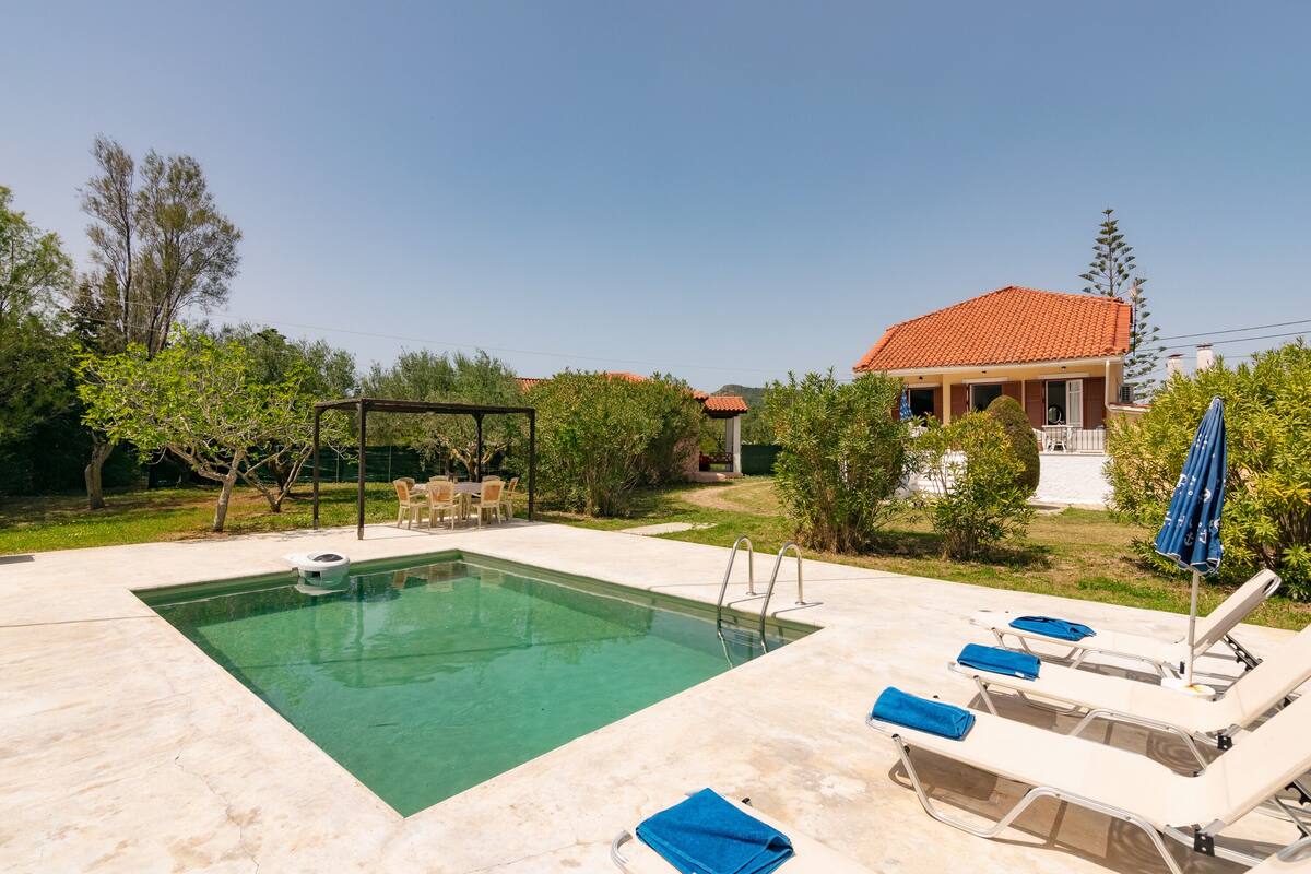 Ein Schwimmbad mit Liegestühlen und einer Pergola, umgeben von Grünflächen und einem Haus mit rotem Dach.