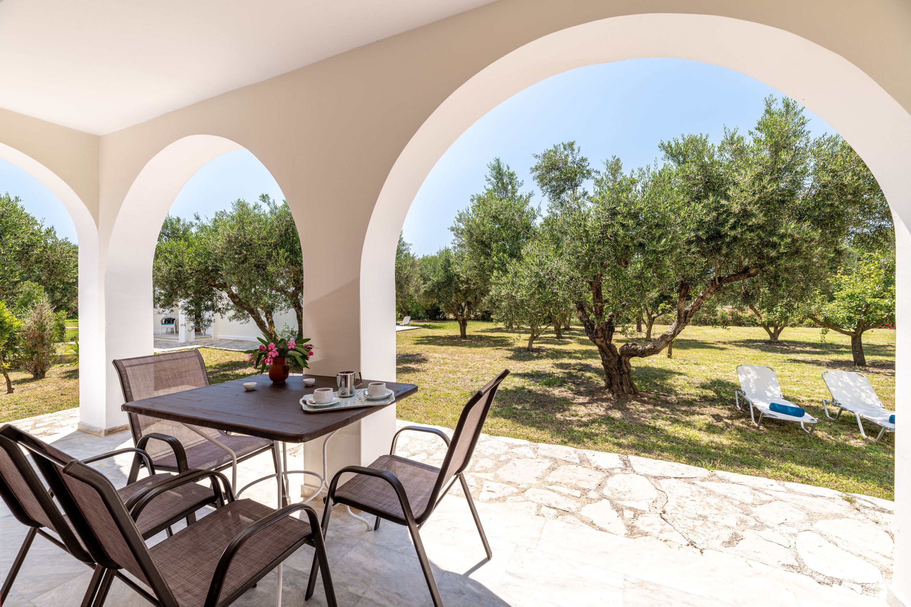 Um pátio externo com mesa e cadeiras, cercado por arcos, com vista para um jardim com árvores e gramado.