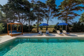 A pool area with clear water, a wooden swing chair, and two lounge chairs under blue umbrellas. - Lenna's Villas (Zakynthos)