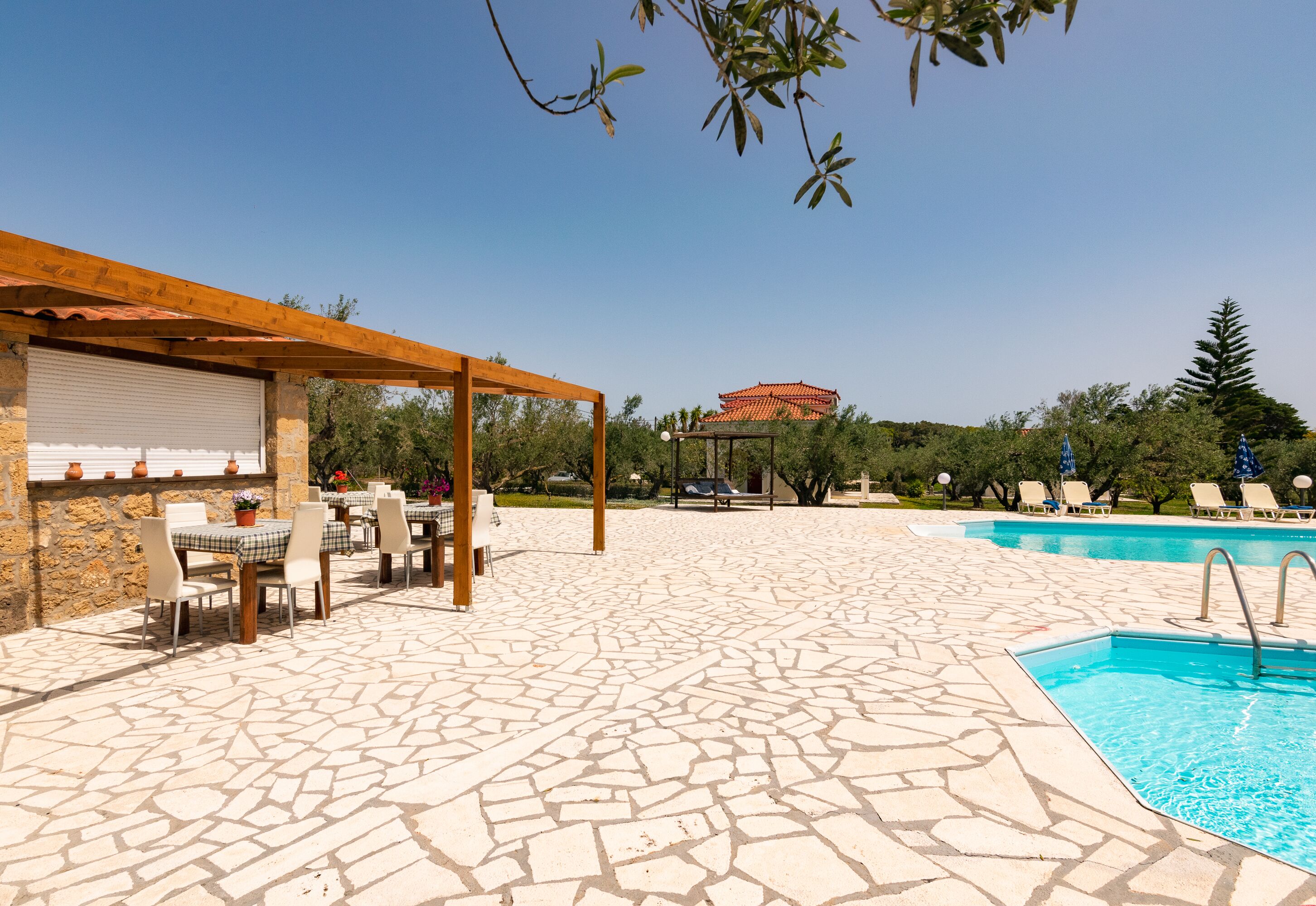 An outdoor pool area with stone flooring, a covered seating area, and a traditional building in the background.