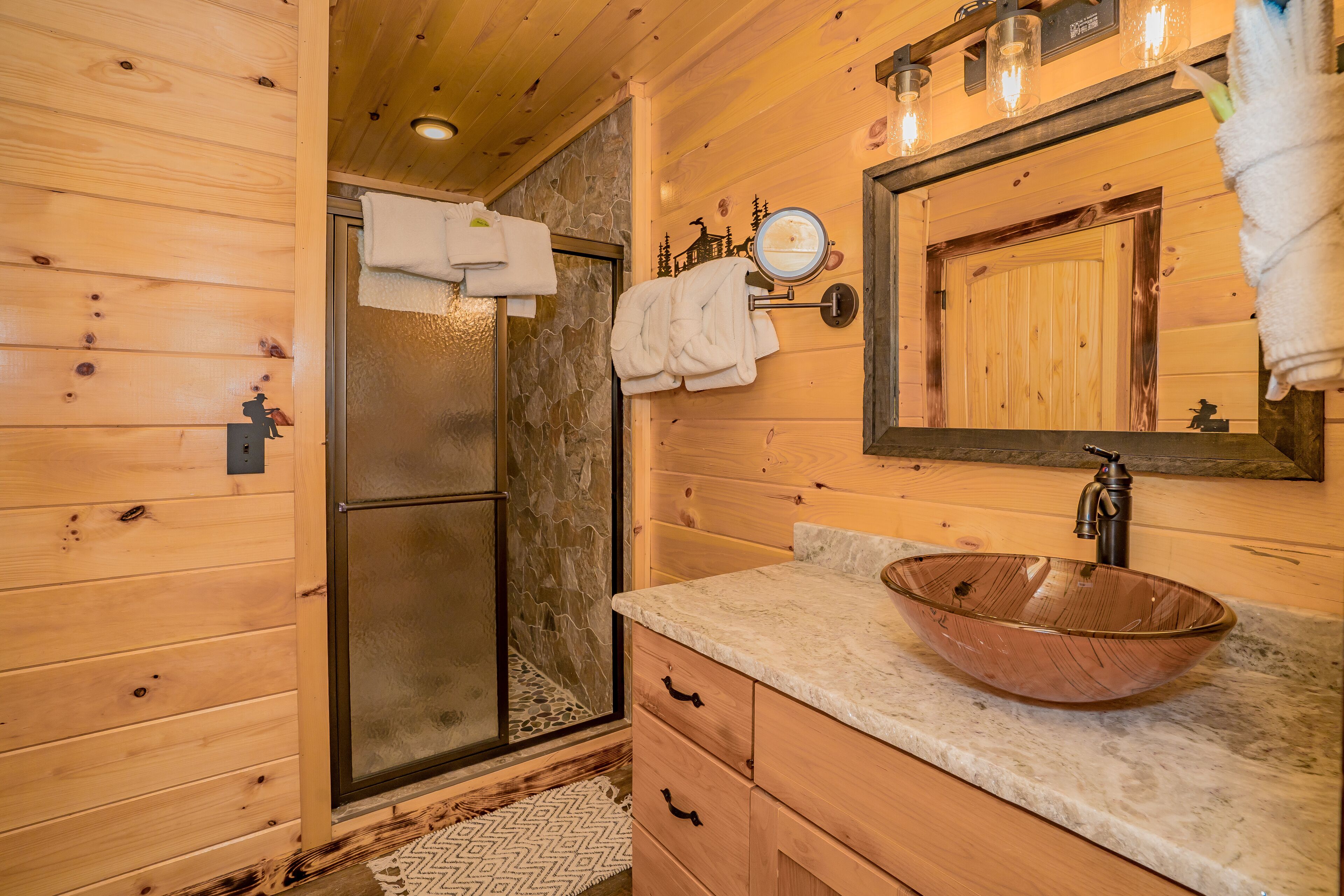 A wooden cabin bathroom with a glass shower door, a wooden vanity with a copper sink, and a mirror above the sink.