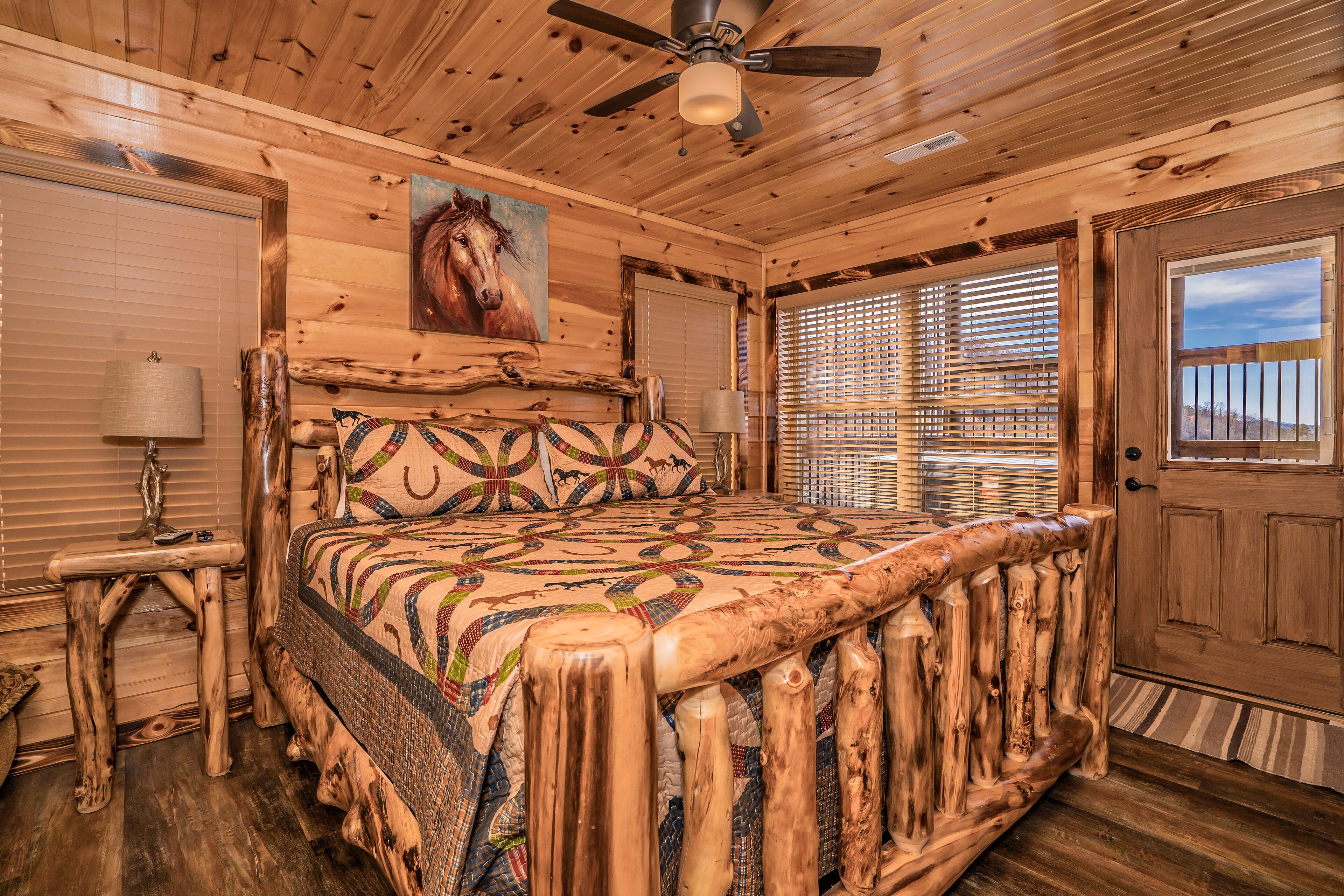 A wooden cabin bedroom with a bed, a ceiling fan, a painting, and a door.