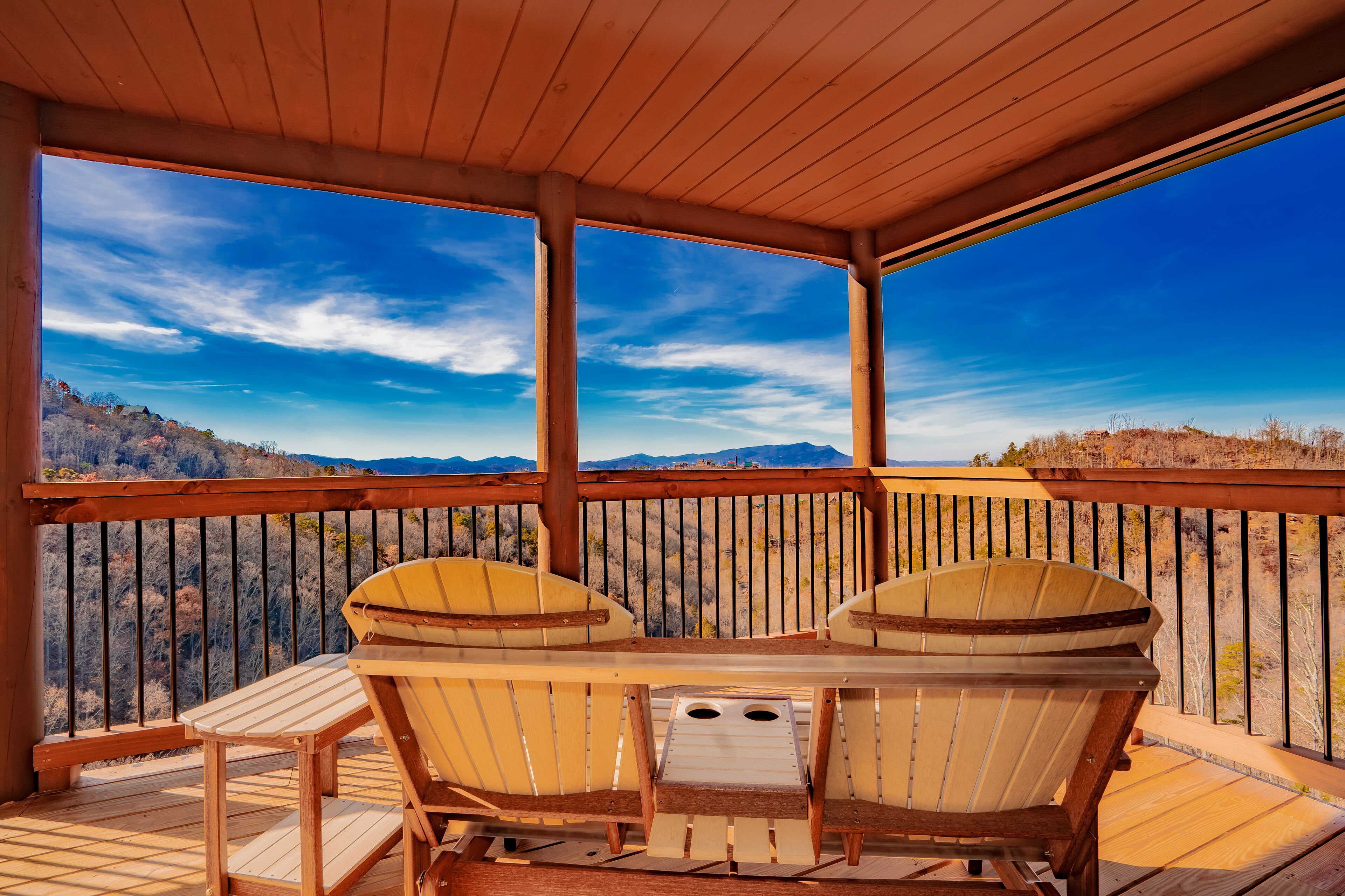 Una terraza de madera con dos sillas Adirondack y una mesa, con vista a un paisaje montañoso.