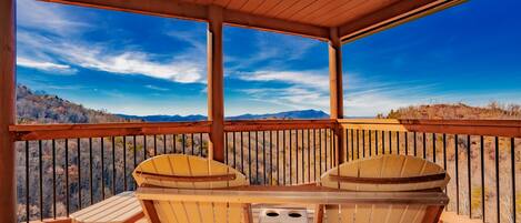 Una terraza de madera con dos sillas Adirondack y una mesa, con vista a un paisaje montañoso.